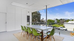 Un'area pranzo moderna con tavolo bianco e sedie verdi, ampie finestre con vista sul quartiere e cielo sereno.