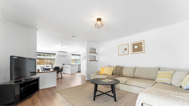 A modern living room with a flat-screen TV, a sofa, a coffee table, and a kitchen in the background. - Trail Side Cottage (Clare)
