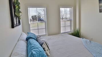 A bedroom with a bed, two windows with blinds, a wall-mounted plant, and a small bedside table.