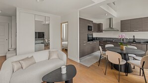 A modern living room with a sofa, a round dining table, and a kitchen area with cabinets and appliances. - Charming Family Duplex in a Peaceful Residence (Cannes)