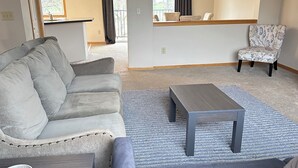 Ein modernes Wohnzimmer mit einem grauen Sofa, einem passenden Sessel, einem Holztisch und einem kleinen Beistelltisch mit einem weißen Gegenstand.