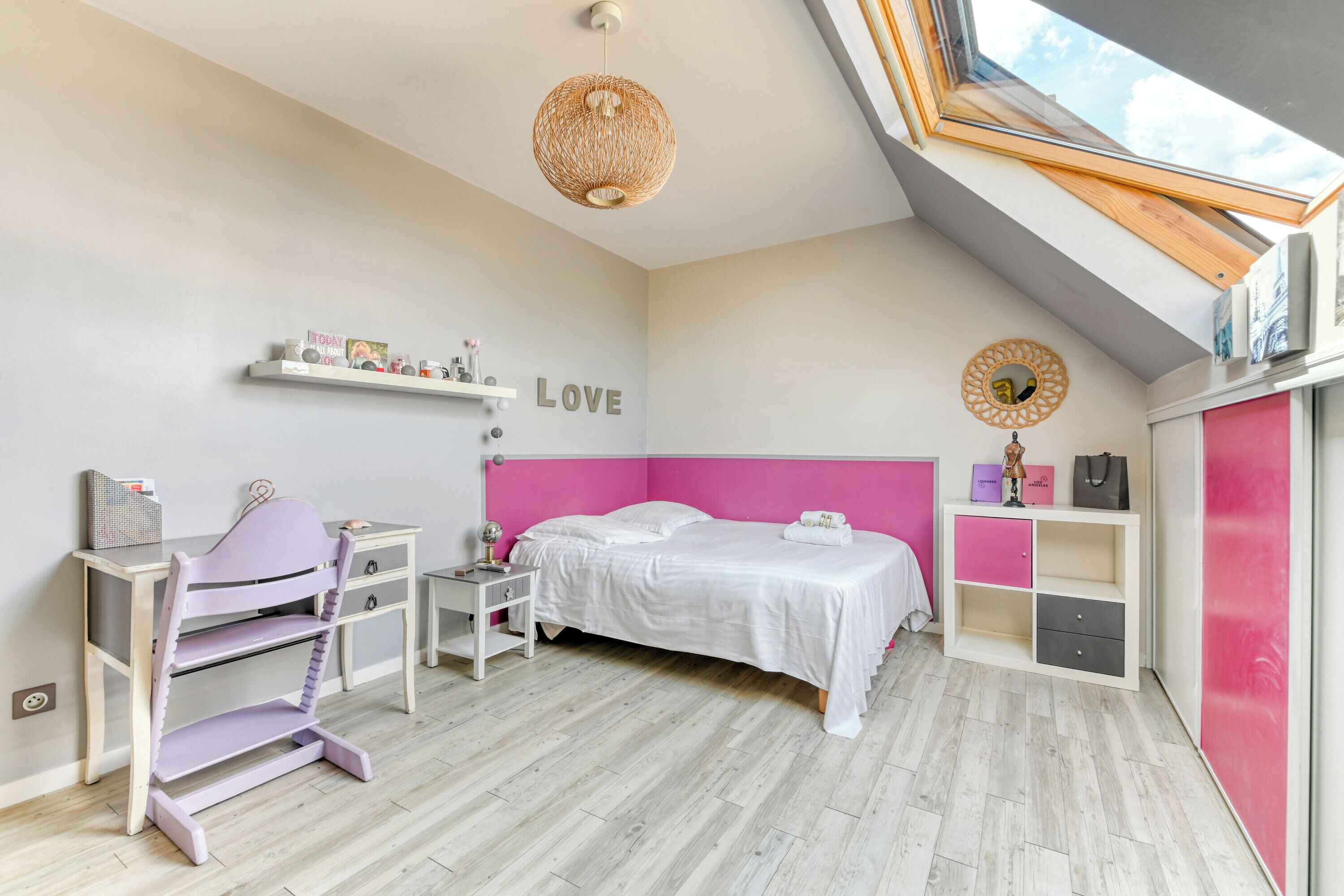 Une chambre à coucher comprenant un lit, un bureau, une chaise, une étagère avec divers objets et une lucarne.