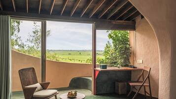 A room with a wooden ceiling, a desk with a chair, a small table with fruit, and a view of a green landscape through large windows.