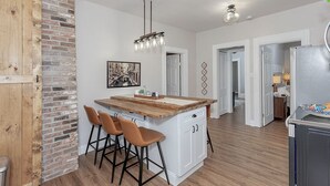 Een keuken met een centraal eiland, houten barstoelen, een hangende lamp en een accentmuur van baksteen.