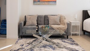 Un salon moderne avec un canapé gris, une table basse en verre et un tableau représentant un coucher de soleil sur l’océan.