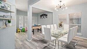 A modern dining area with a glass table and white chairs, a foosball table, and a 'Welcome' sign on a shelf. - Stylish Modern House  4BR/3BA  Home in McKinney (McKinney)