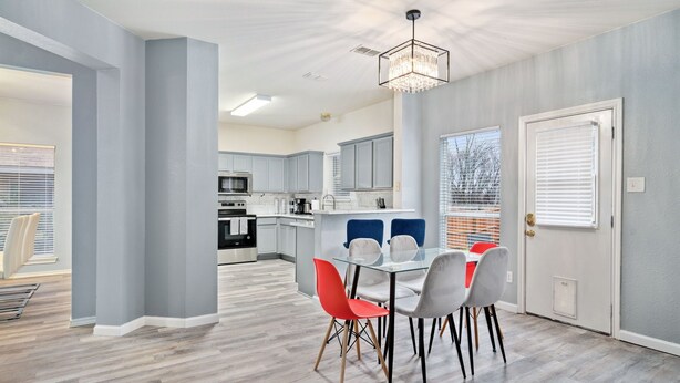 A modern kitchen with grey cabinets, a central island, and a dining area with a glass table and red chairs. - Stylish Modern House  4BR/3BA  Home in McKinney (McKinney)