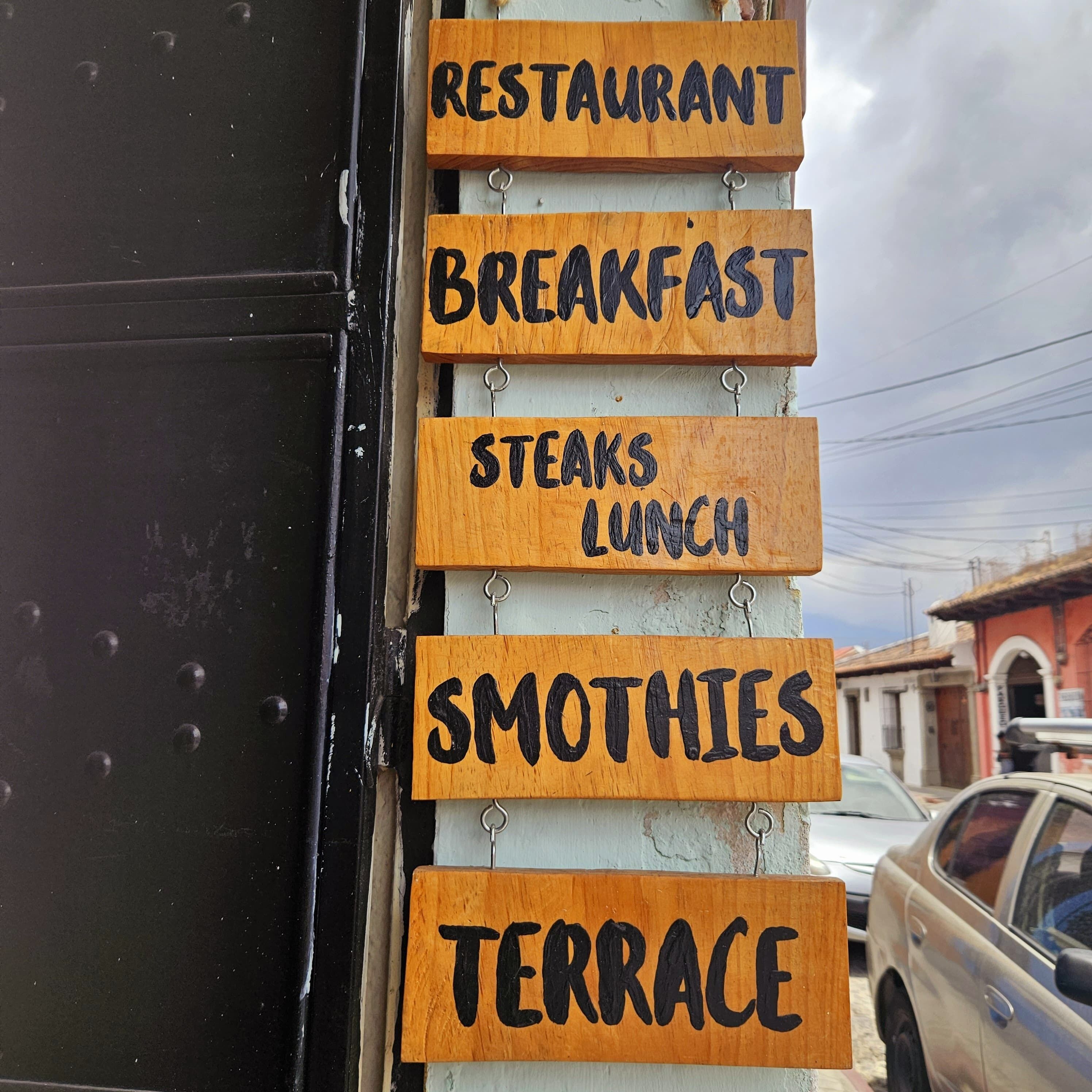 Un panneau vertical en bois, avec des indications pour les différents types de repas proposés : restaurant, petit-déjeuner, déjeuner avec plats de viande, smoothies et terrasse.
