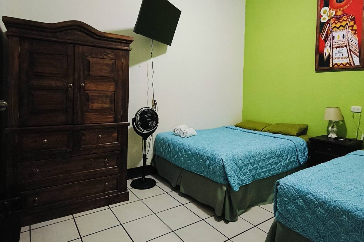 Ein Zimmer mit zwei Betten, einem Holzschrank, einem Standventilator und einem an der Wand befestigten Fernseher.