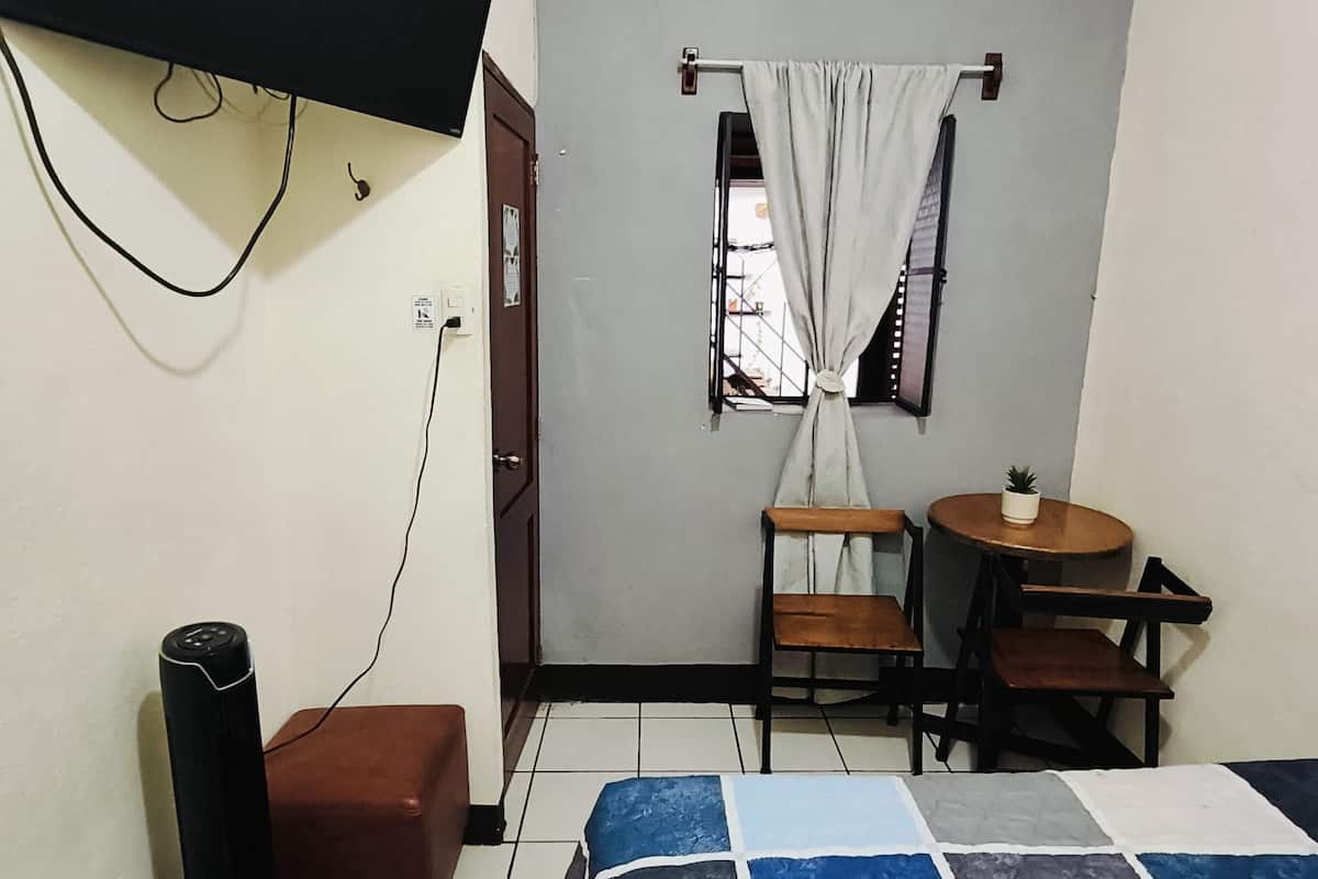 Ein kleiner, sauberer Raum mit einem Bett, einem an der Wand befestigten Fernseher, einem Schreibtisch mit Stuhl und einem Fenster mit Vorhängen.
