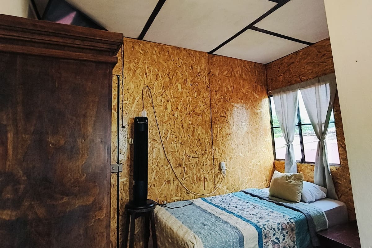 Ein Zimmer mit Holzboden, einem Bett mit blauen und weißen Kissen, einem Holzschrank und einem kleinen Tisch mit einer Lampe.