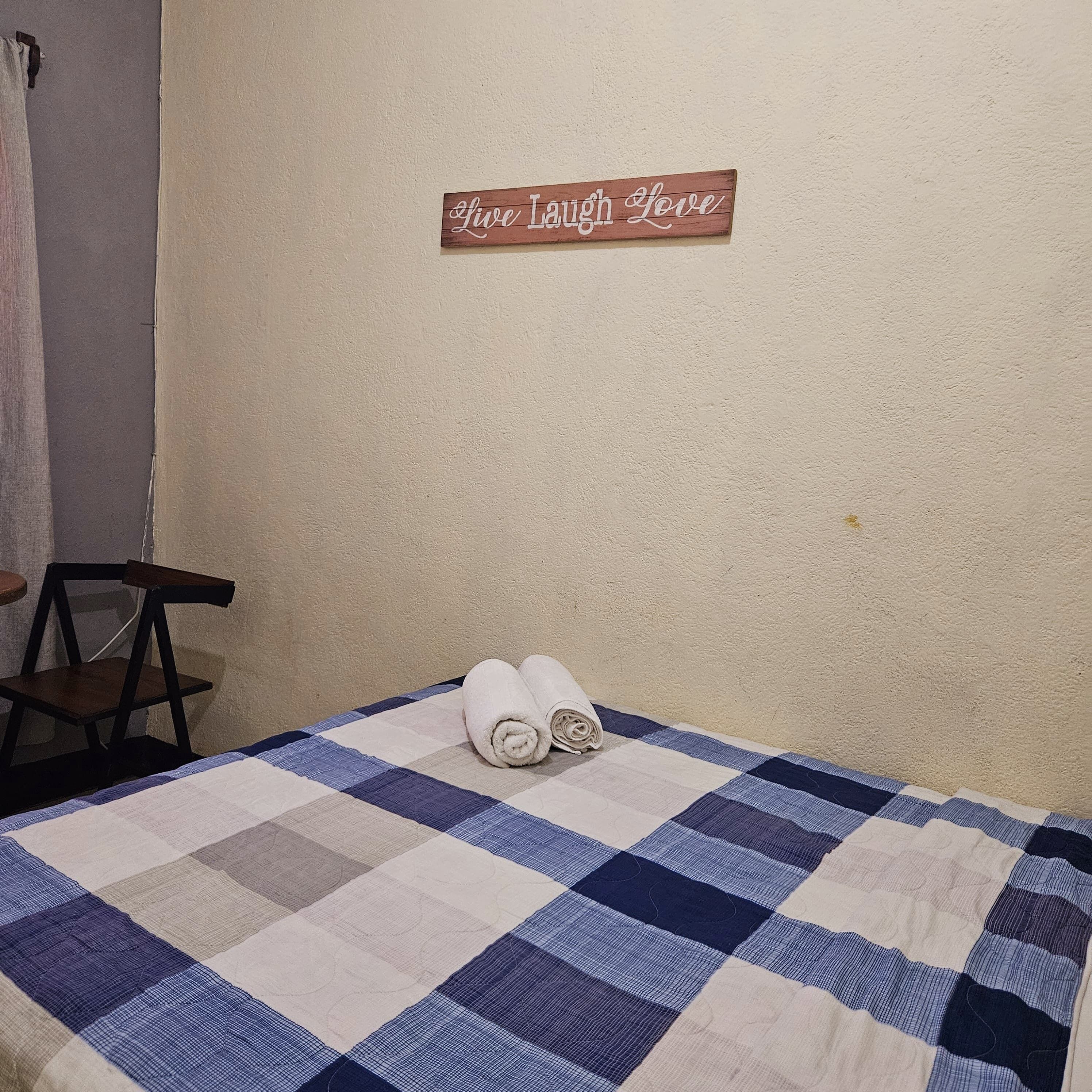 Ein Zimmer mit einem Bett, das mit einer blau-weiß karierten Decke bezogen ist, einem aufgerollten weißen Handtuch auf dem Bett, einem Holzschild an der Wand und einem Stuhl, der an der Wand lehnt.
