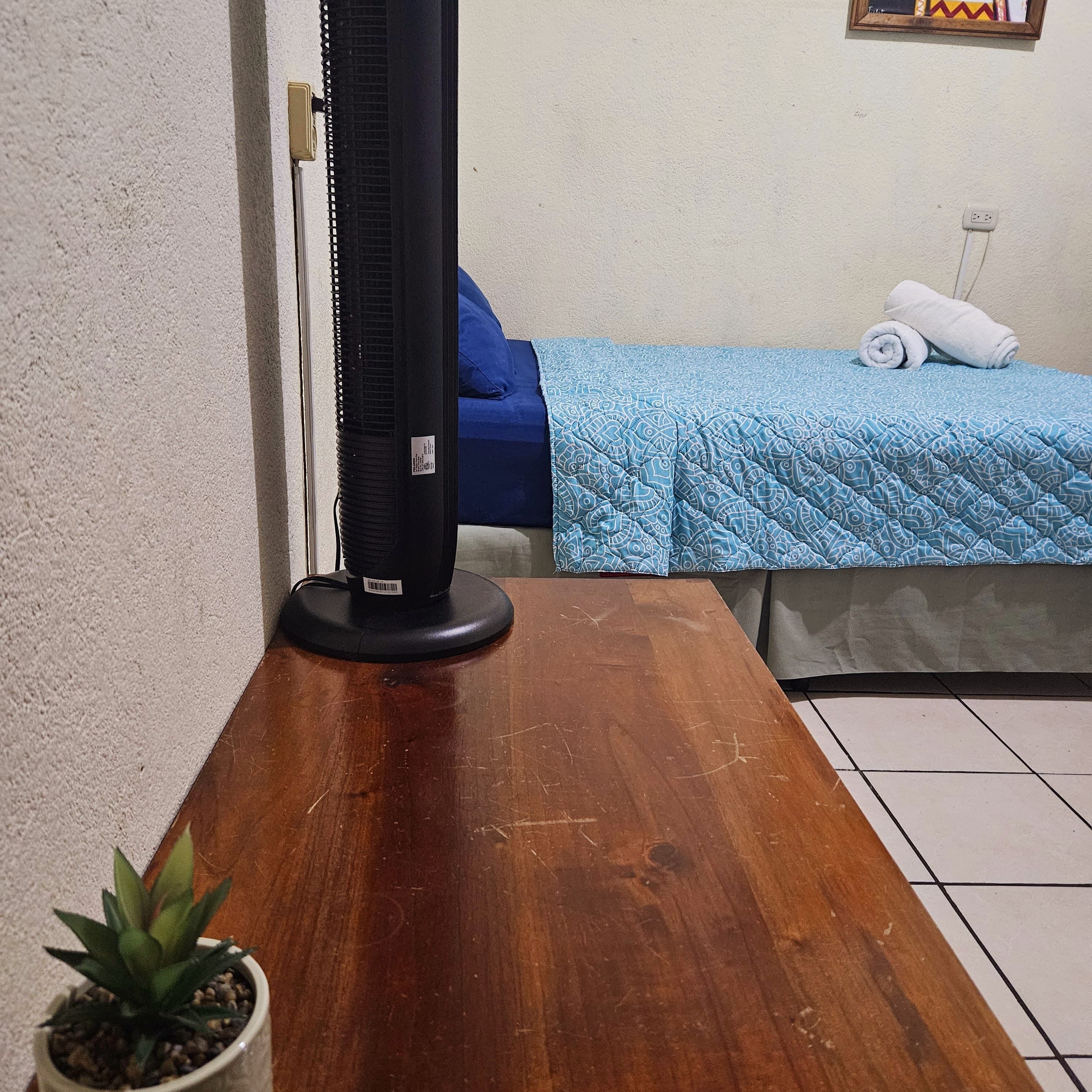 Une pièce avec une table en bois, un ventilateur sur pied, un lit recouvert d’une couette bleue et une petite plante en pot.