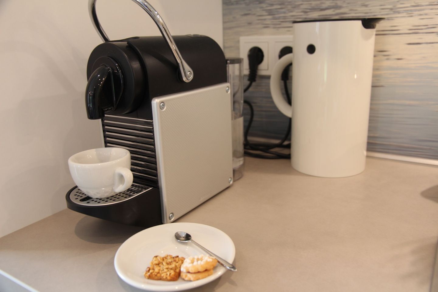 A coffee machine with a cup holder, a white mug, and a plate with cookies and a spoon on a countertop.
