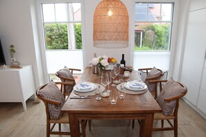 Een eetgedeelte met een houten tafel voor zes personen, omringd door stoelen met kussens. Daarboven hangt een geweven hanglamp, en de kamer heeft grote ramen met uitzicht op groen buiten.