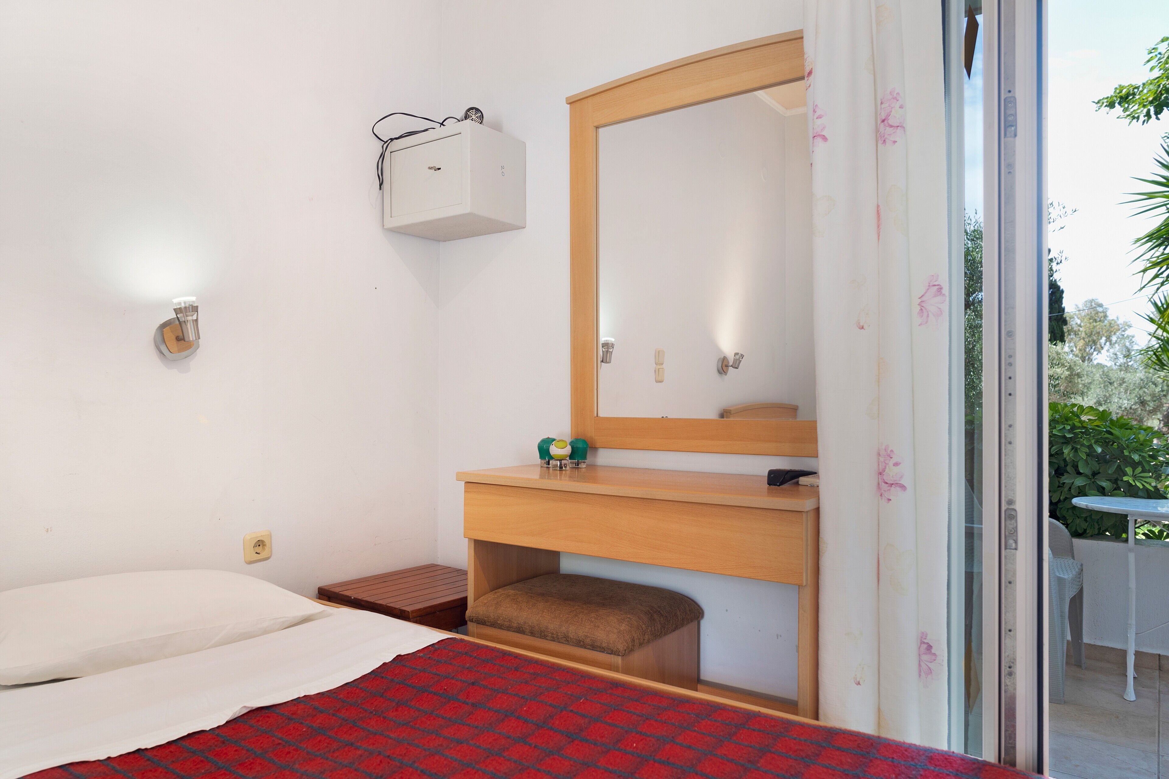 A bedroom with a bed, a wooden bedside table, a small bench, a mirror, and a wall-mounted electrical box.