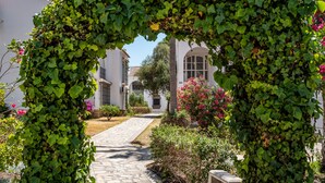 A pathway leading through a lush garden with a vine-covered archway, surrounded by white buildings with arched windows and a clear sky. - Apartment 'M - 1' with Shared Pool, Private Terrace and Wi-Fi (Torrox costa)