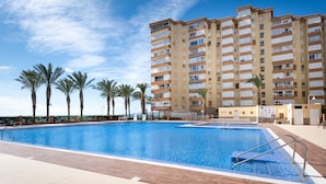 A swimming pool with a wooden deck, palm trees, and a tall building in the background. - Apartment 'M - 1' with Shared Pool, Private Terrace and Wi-Fi (Torrox costa)