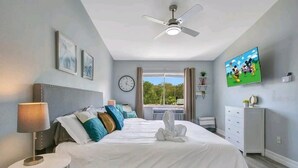 A hotel room with a bed, a bedside table, a lamp, a ceiling fan, a clock, a TV, and a window with a view of greenery.