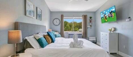 A hotel room with a bed, a bedside table, a lamp, a ceiling fan, a clock, a TV, and a window with a view of greenery.