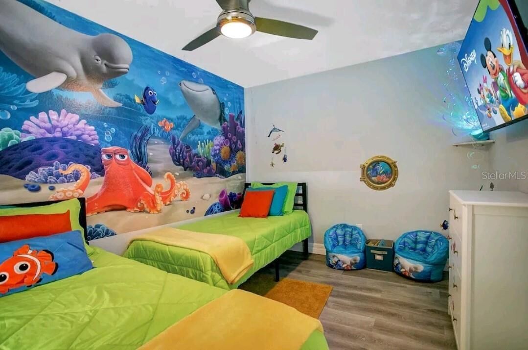 Habitación infantil con dos camas, ropa de cama colorida, un mural de una escena submarina, un ventilador de techo y un televisor con temática de Disney.