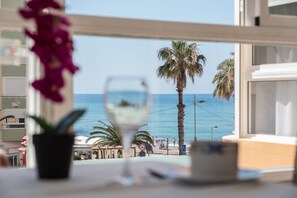 Une vue depuis une fenêtre donnant sur une plage bordée de palmiers, un verre de vin blanc et une table sur laquelle est posée une plante en pot.