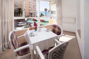 Un balcon avec une table dressée pour deux personnes, des chaises avec des coussins à carreaux, une vue sur la plage et un bâtiment en arrière-plan.