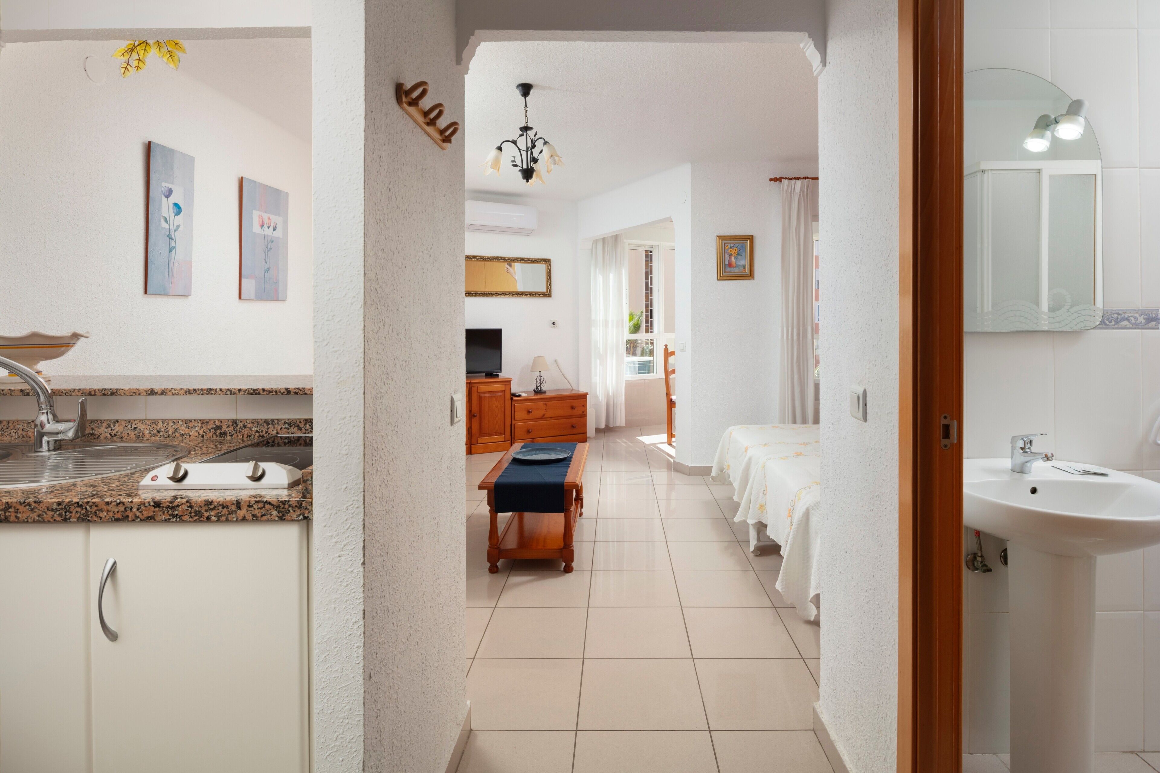 Une chambre d’hôtel équipée d’une kitchenette, d’un coin repas, d’une salle de bain avec un lavabo et un miroir, et d’un couloir menant à une autre chambre.
