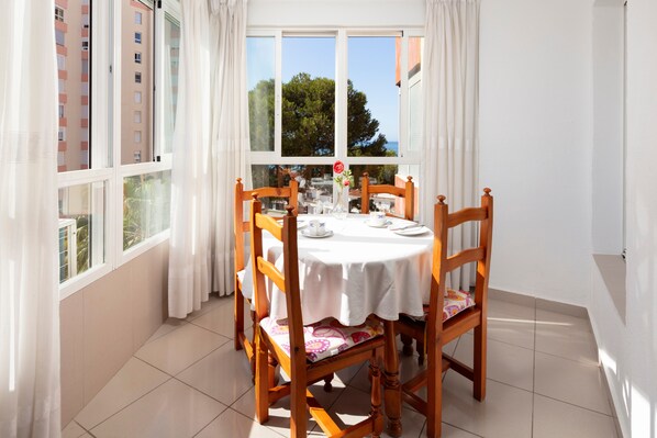 A dining area with a round table, chairs, a vase with flowers, and a view through large windows.