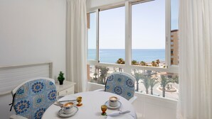 Un comedor con una mesa preparada para dos personas, sillas con tapicería estampada, vista al mar y una ventana con cortinas blancas.