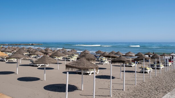 Una playa con hileras de sombrillas de paja y sillas reclinables blancas.