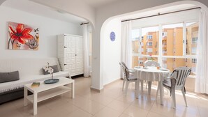 Un salon comprenant un canapé, une table basse, une table à manger avec des chaises, un meuble et une grande fenêtre donnant sur des immeubles.