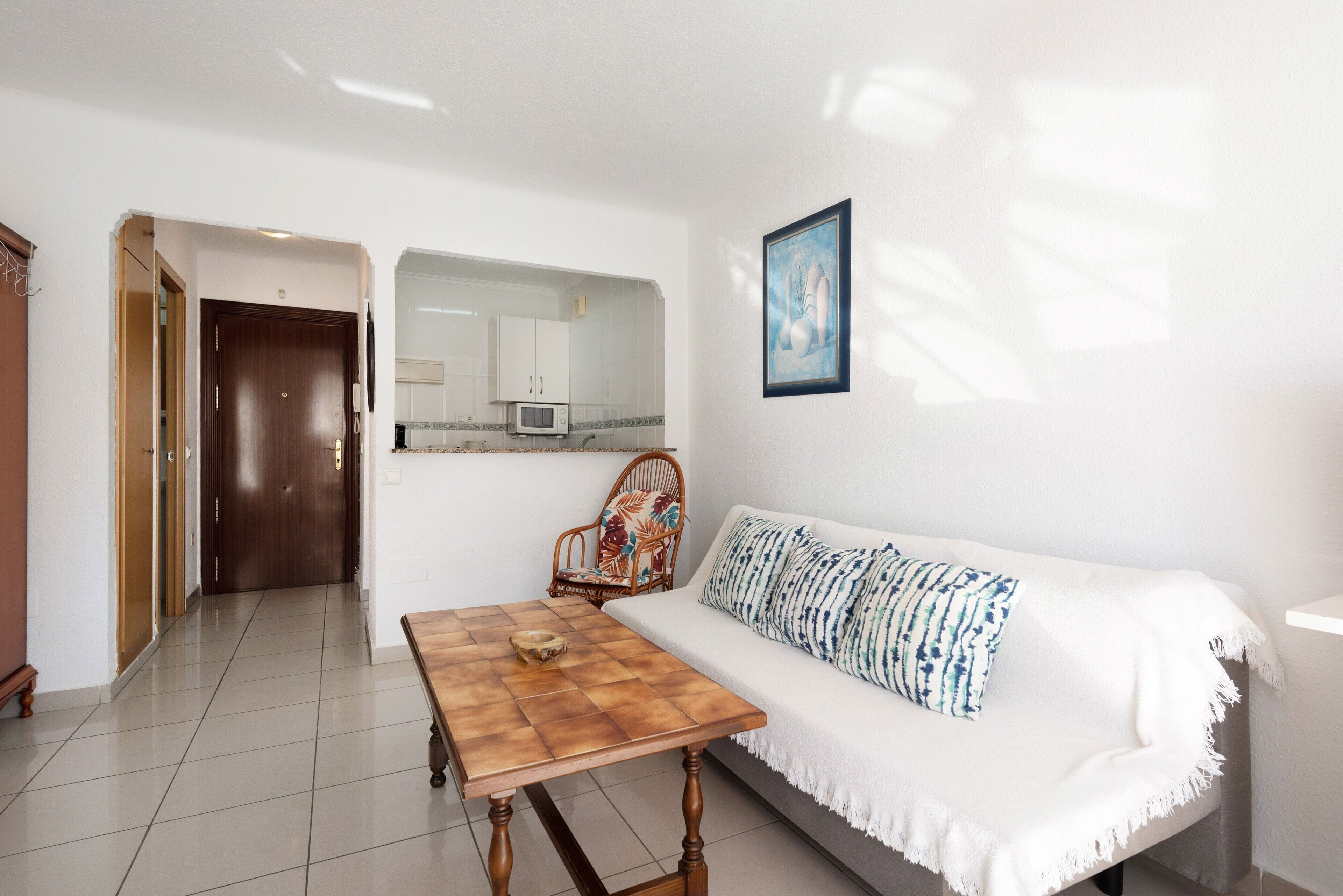 Un salon avec un canapé blanc, une table basse en bois, un fauteuil à motifs, un tableau encadré au mur et, au fond, un coin cuisine.