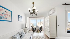 Une pièce avec un canapé blanc, une table à motifs, un lustre, une vue par la fenêtre et une armoire blanche.