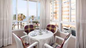 A balcony with a table set for two, white chairs with plaid cushions, and a view of the sea and buildings.