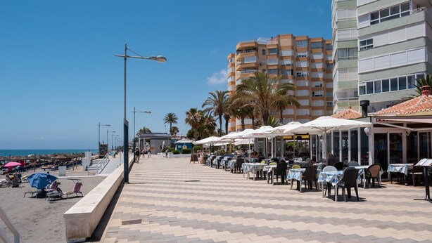 A seaside promenade with outdoor seating, palm trees, and a tall building in the background.