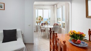 A room with a bed, a dining table with chairs, a view of the sea, and a window with curtains.