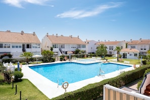 Una piscina rodeada de edificios blancos con tejados de tejas rojas, un cielo azul despejado y vegetación bien cuidada.