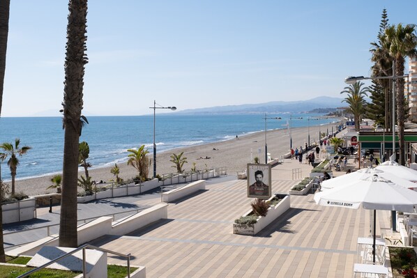 A coastal promenade with a beach, palm trees, and outdoor seating areas.