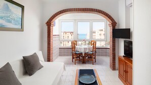 Une pièce avec un canapé blanc, une table à manger avec des chaises, un téléviseur et une vue sur la mer par une fenêtre en arc de cercle.