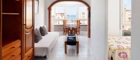 A bedroom with a bed, a wooden wardrobe, a dining table with chairs, and a balcony with a view of the sea.