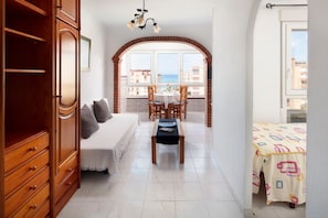 A bedroom with a bed, a wooden wardrobe, a dining table with chairs, and a balcony with a view of the sea.
