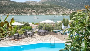 A swimming pool with lounge chairs and umbrellas, a stone deck, and a view of a lake with boats and mountains in the background. - Holiday Home 'Villa 2' with Sea View, Wi-Fi and Air Conditioning (Leykada)