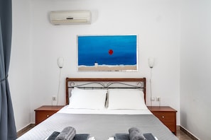 A bedroom with a bed, two bedside tables, a wall-mounted air conditioner, and a painting of a beach scene.