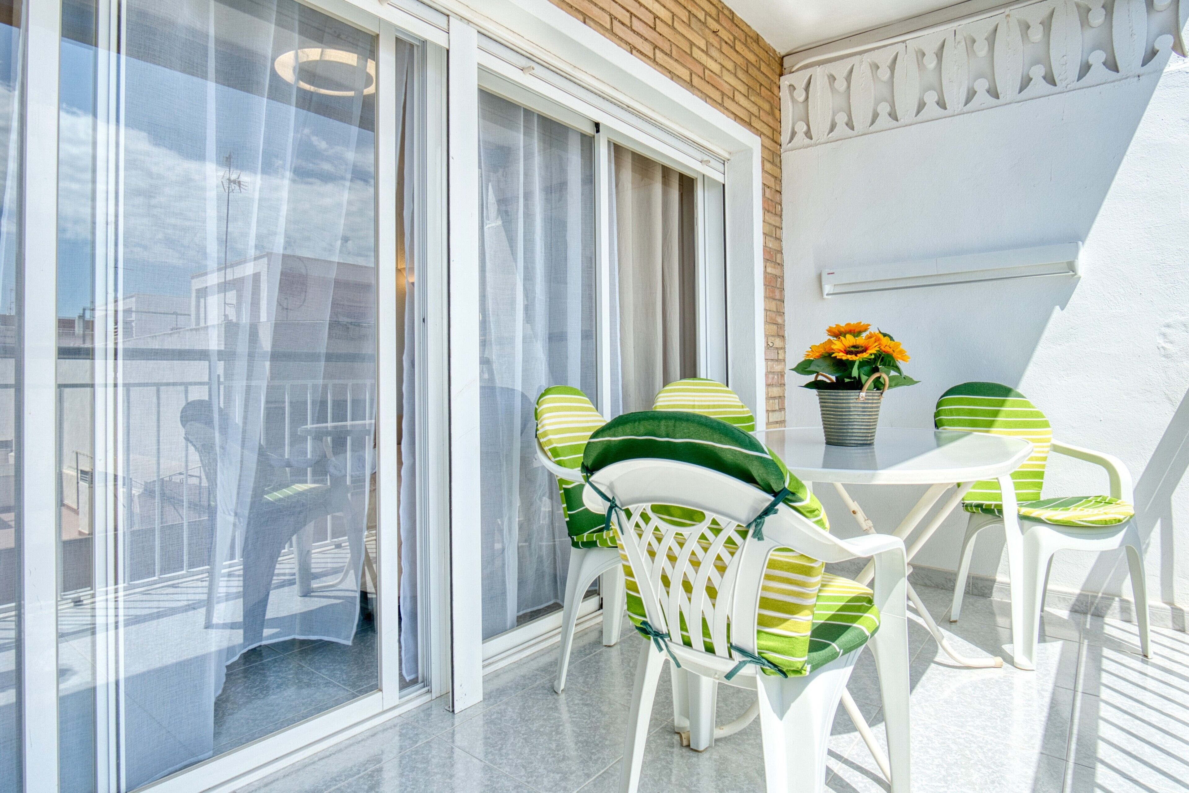 Un balcon avec des chaises blanches et une table, une plante en pot, et une vue à travers une porte vitrée.