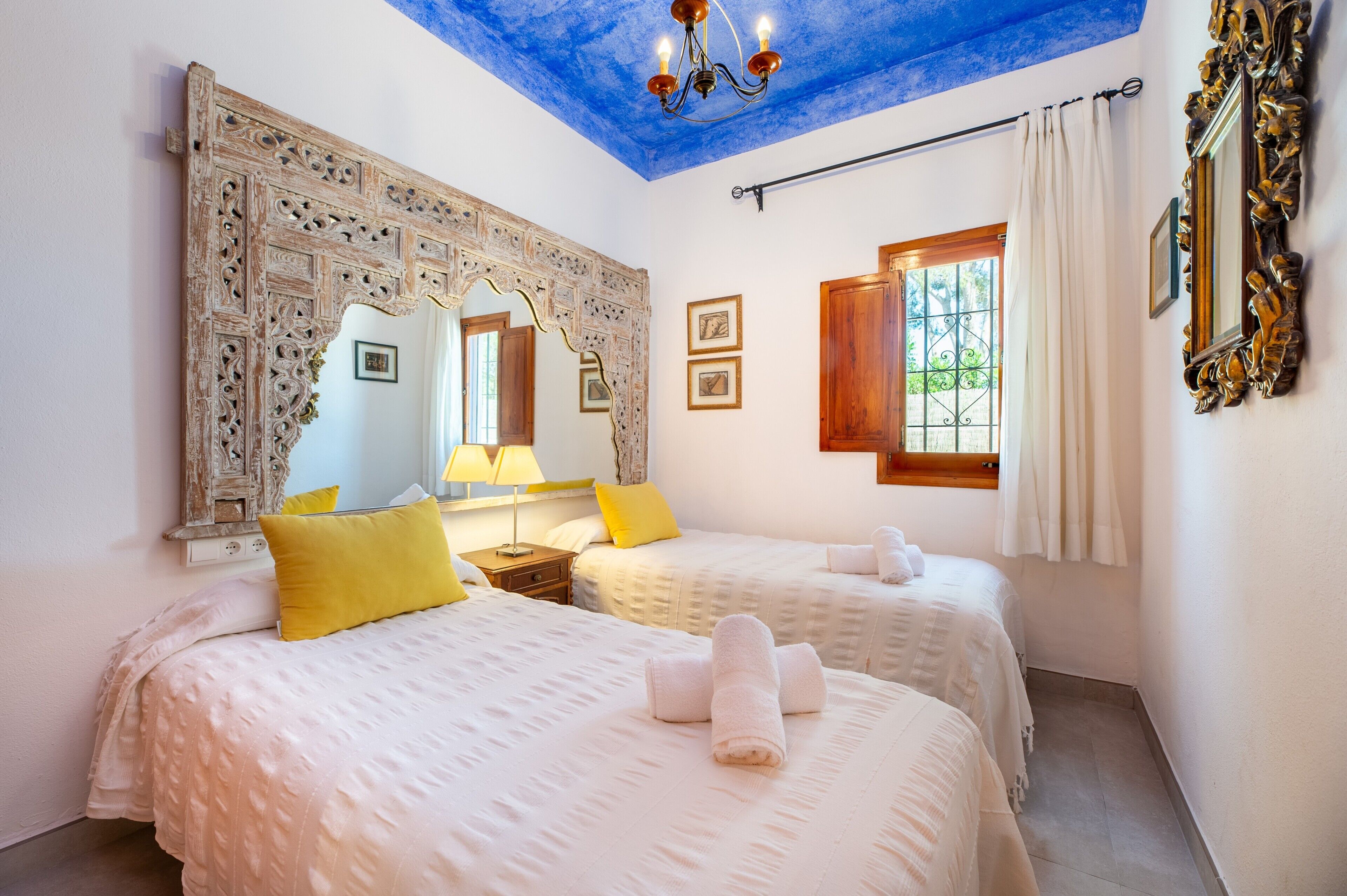 A room with two beds, a chandelier, a large mirror, and a window with curtains.