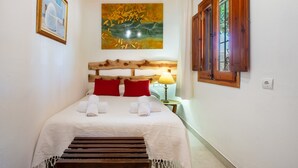 Un lit bien fait, avec du linge de lit blanc et des oreillers rouges, une tête de lit en bois, un tableau au-dessus du lit et une fenêtre avec des volets en bois.