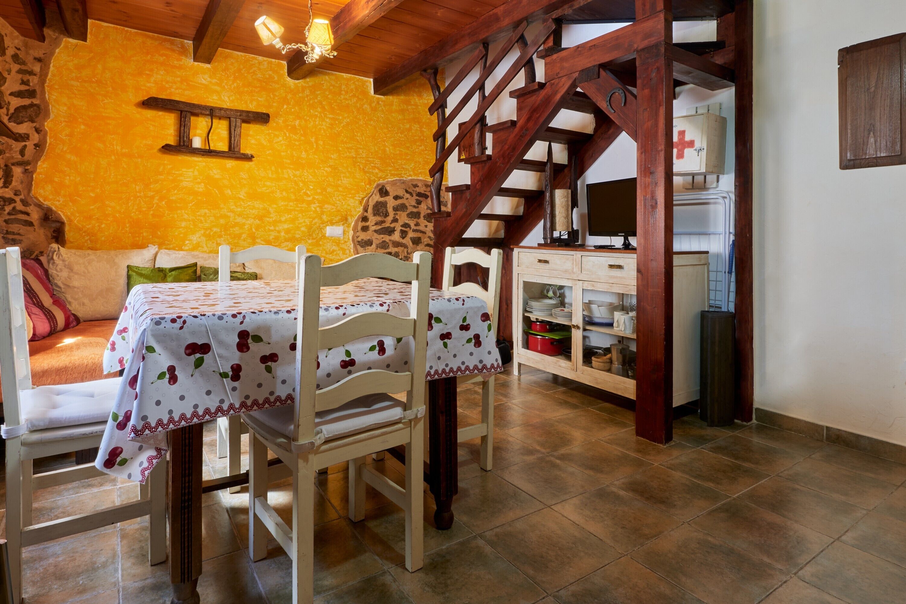 A dining area with a table set for two, wooden chairs, a stone wall, a staircase, and a kitchen area with cabinets and a refrigerator.
