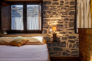 Ein Schlafzimmer mit Steinwand, einem Bett mit weißer Bettwäsche, einer Holzkommode und einem Fenster mit Vorhängen.
