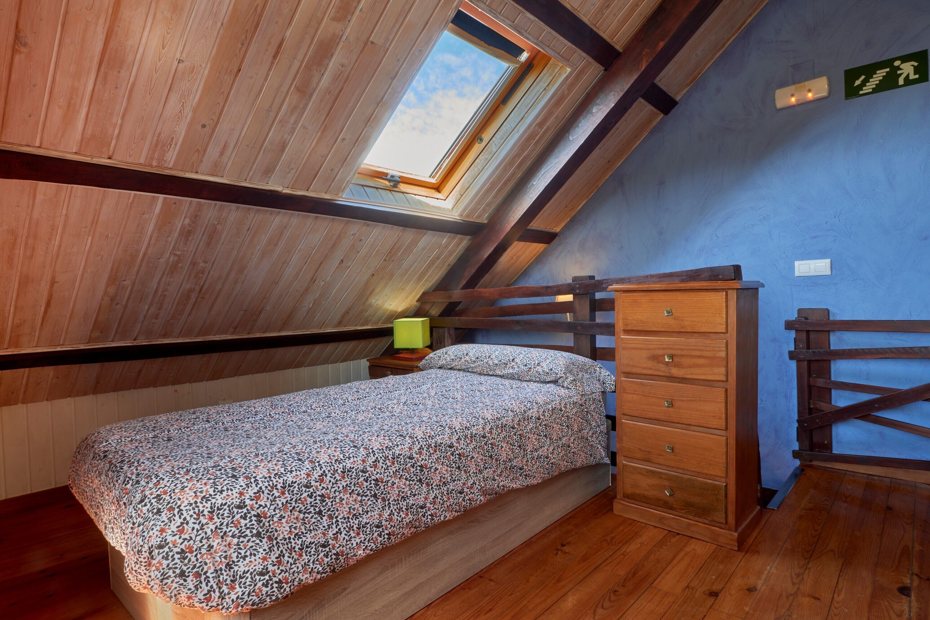Un dormitorio con techo inclinado, una cama con colcha estampada, un aparador de madera y un tragaluz.